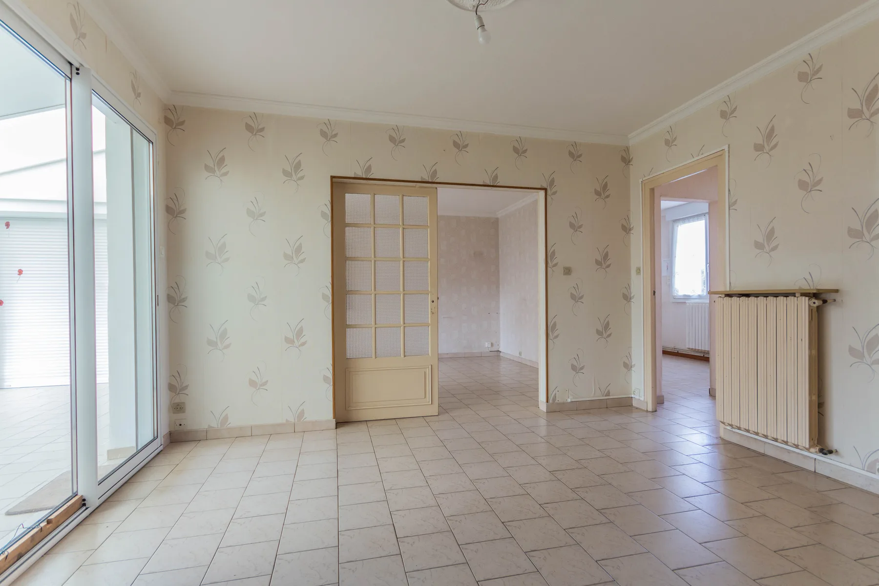 Pièce de vie avant rénovation avec carrelage, papier peint, baie vitrée et radiateur